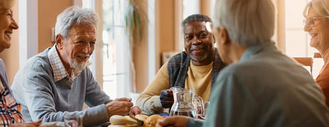 A group of elderly came home residents take part in a group activity. They are smiling and sat around a table with refreshments.