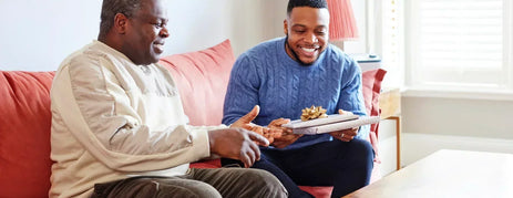 Older man with dementia on a coral sofa opening a wrapped Father's Day gift from his son