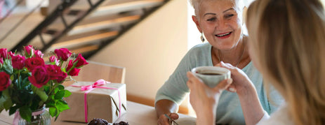 A daughter and mother sitting at a table drinking coffee with a gift, a bunch of roses and chocolate muffins on the table.