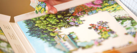 A close-up of a person's hands placing a large piece into a carefully designed dementia jigsaw puzzle of a vibrant garden scene. 