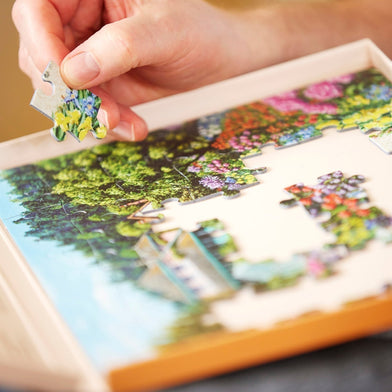 A close-up of a person's hands placing a large piece into a carefully designed dementia jigsaw puzzle of a vibrant garden scene. 
