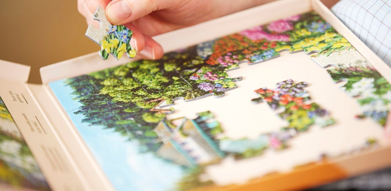 A close-up of a person's hands placing a large piece into a carefully designed dementia jigsaw puzzle of a vibrant garden scene. 