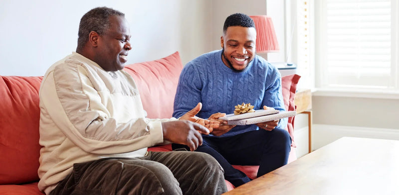 Dad with dementia on a coral sofa opening a wrapped Father's Day gift from his son