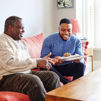 Older man with dementia on a coral sofa opening a wrapped Father's Day gift from his son 