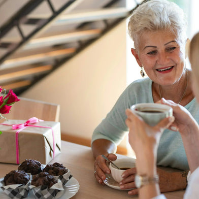 A daughter and mother sitting at a table drinking coffee with a gift, a bunch of roses and chocolate muffins on the table.