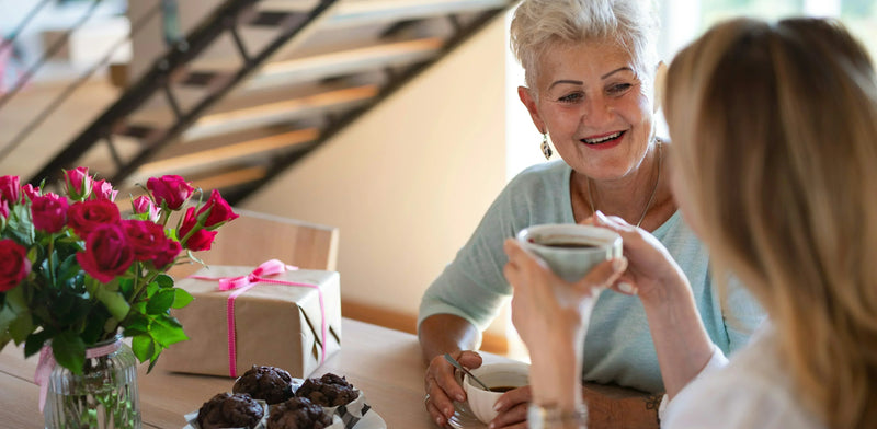 A daughter and mother sitting at a table drinking coffee with a gift, a bunch of roses and chocolate muffins on the table.