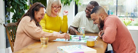 A happy group sitting at a wooden table playing Snakes and Ladders board game adapted for dementia