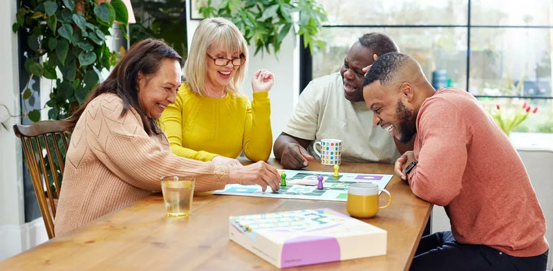 A happy group sitting at a wooden table playing Snakes and Ladders board game adapted for dementia