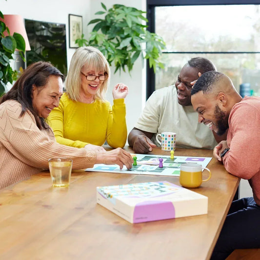 A happy group sitting at a wooden table playing Snakes and Ladders board game adapted for dementia